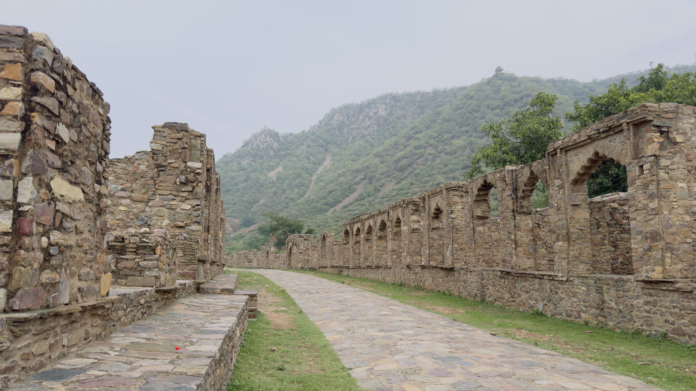Ruined stone shopfronts with arched niches lining a paved street at Bhangarh, green Aravalli hills behind