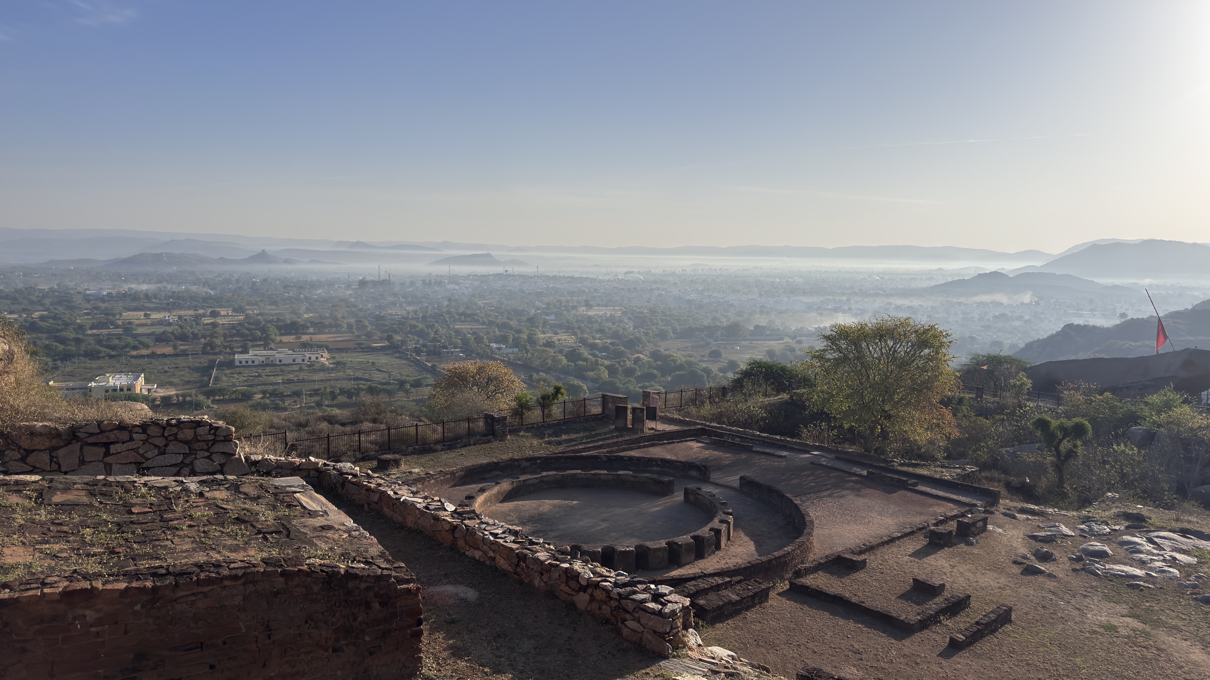 Circular stone foundation of the Buddhist monastery ruins on the hilltop at Bijak ki Pahadi, with a hazy green plain and low hills below